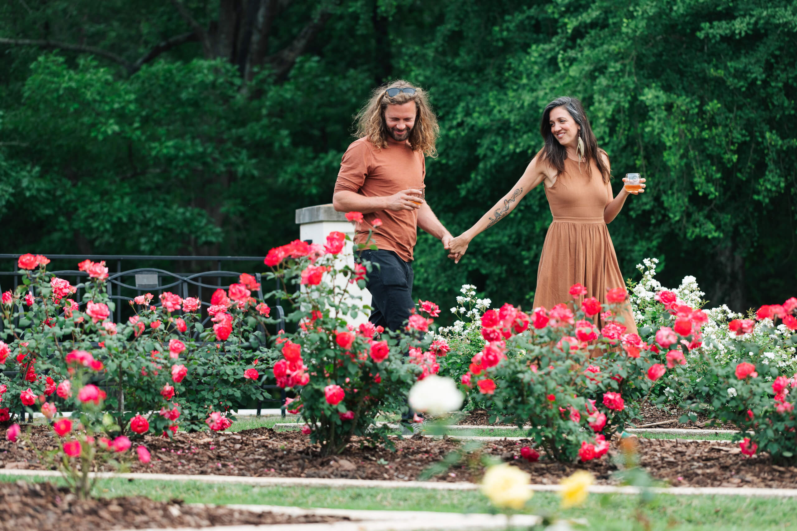 Woman holding hands with man as they walk past rose garden.